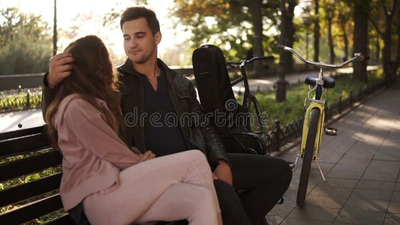 Ένα όμορφο κορίτσι με τη μακριά καφετιά τρίχα κάθεται το αγκάλιασμα με τον όμορφο τύπο brunette της σε έναν πάγκο πάρκων Πορτρέτο