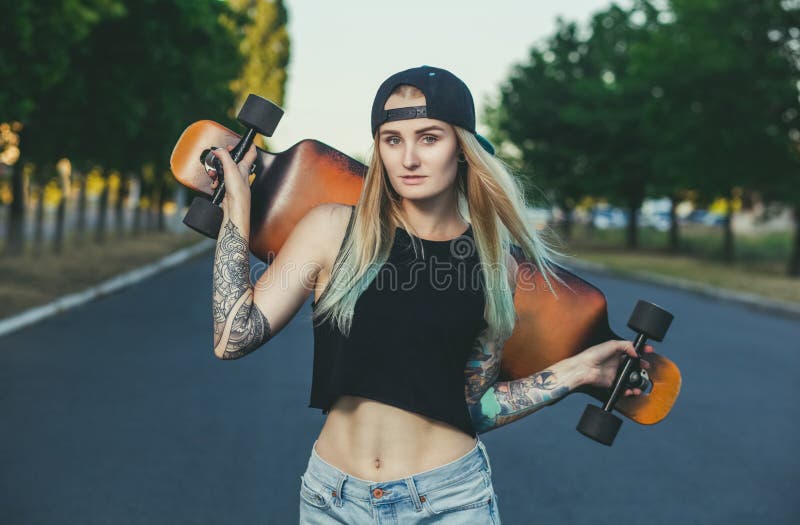 Ένα όμορφο, προκλητικό hipster ξανθό με την μπλε τρίχα στη δερματοστιξία στέκεται με ένα longboard στοκ φωτογραφία με δικαίωμα ελεύθερης χρήσης Ένα όμορφο, προκλητικό hipster ξανθό με την μπλε τρίχα στη δερματοστιξία στέκεται με ένα longboard στοκ φωτογραφία με δικαίωμα ελεύθερης χρήσης