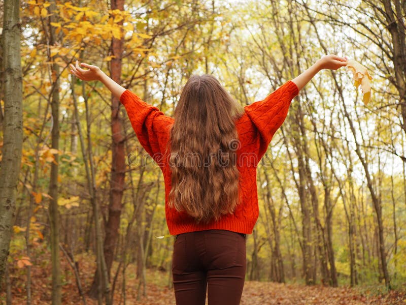 A teenager lifting her hands up with joy while in the forest. A girl wearing orange sweater and brown jeans with long hair on walk in an autumn forest. A teenager lifting her hands up with joy while in the forest. A girl wearing orange sweater and brown jeans with long hair on walk in an autumn forest.