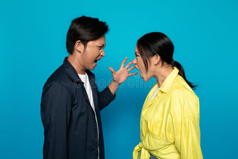 An Asian millennial man and woman are facing each other with open mouths, as if engaged in a heated argument or shouting match, against a solid turquoise background, studio. An Asian millennial man and woman are facing each other with open mouths, as if engaged in a heated argument or shouting match, against a solid turquoise background, studio