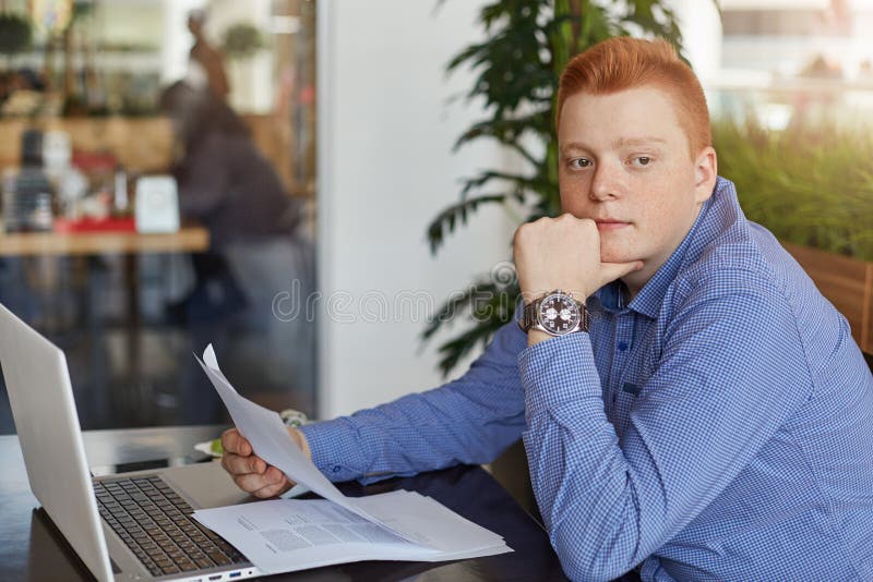 Ένας σκεπτικός νέος redhead επιχειρηματίας που φορά την επίσημη συνεδρίαση πουκάμισων στον πίνακα με το lap-top και τα έγγραφα στ στοκ εικόνες Ένας σκεπτικός νέος redhead επιχειρηματίας που φορά την επίσημη συνεδρίαση πουκάμισων στον πίνακα με το lap-top και τα έγγραφα στ στοκ εικόνες