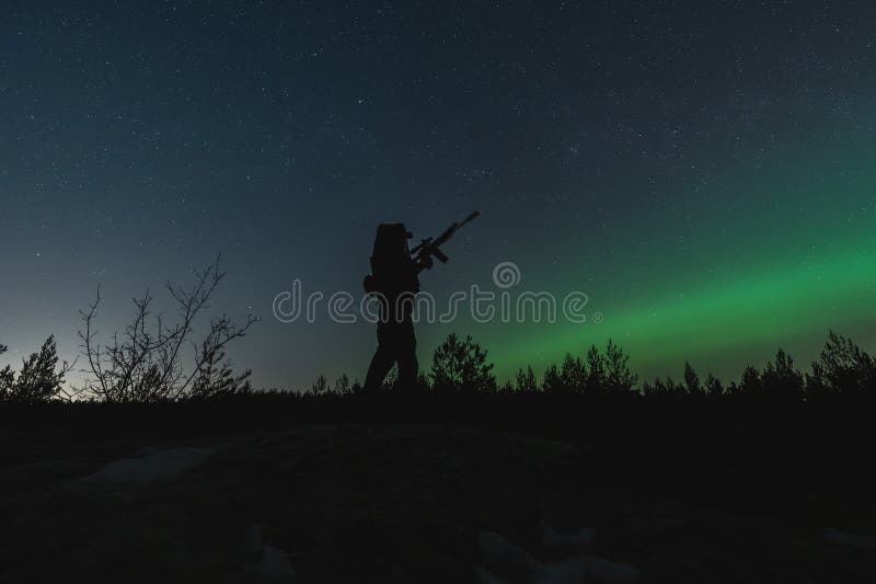 A private military soldier with a night vision device and a rifle with a suppressor in the forest against the background of the starry sky and northern lights. A private military soldier with a night vision device and a rifle with a suppressor in the forest against the background of the starry sky and northern lights.