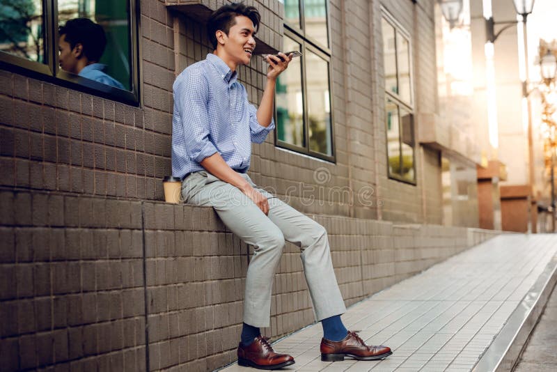 A Smiling Businessman Using Voice Control via Smartphone. Happy Young Man holding a Mobile Phone in the Urban City. A Smiling Businessman Using Voice Control via Smartphone. Happy Young Man holding a Mobile Phone in the Urban City