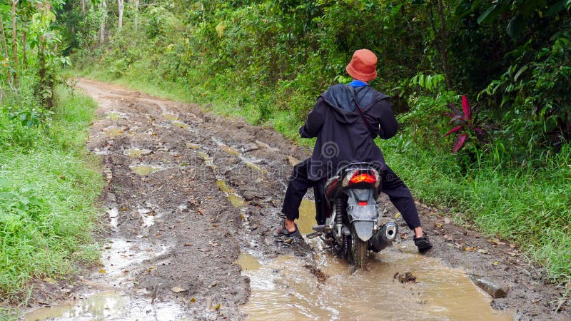 A villager rides a motorbike through a muddy forest road with water and puddles in the village. A villager rides a motorbike through a muddy forest road with water and puddles in the village