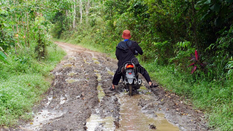 A villager rides a motorbike through a muddy forest road with water and puddles in the village. A villager rides a motorbike through a muddy forest road with water and puddles in the village