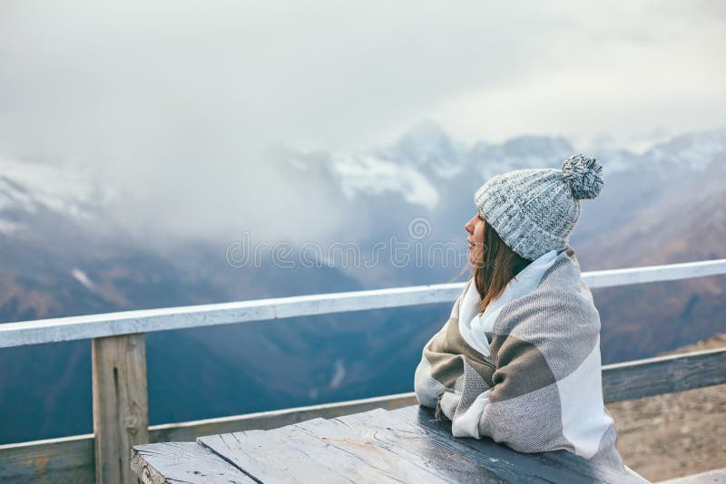 Teen girl wrapped in plaid blanket sitting by table on mountain top with a view. Cold weather, snow on hills. Winter hiking scene. Teen girl wrapped in plaid blanket sitting by table on mountain top with a view. Cold weather, snow on hills. Winter hiking scene