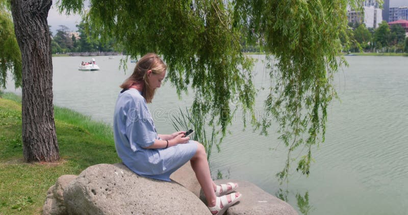 έφηβος που κάνει παρέα στο smartphone ενώ κάθεται δίπλα στη λίμνη στο πάρκο