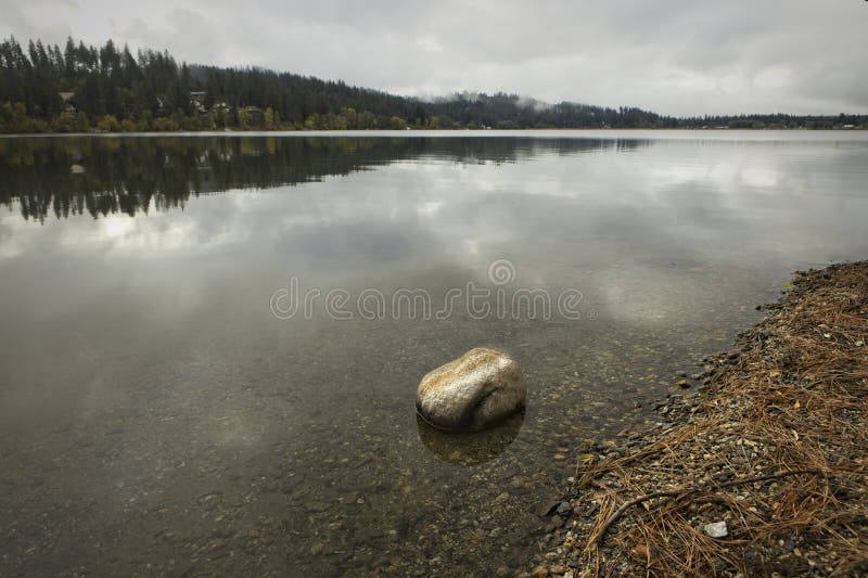 A small boulder is in the calm water of Hauser Lake under a cloudy sky in north Idaho. A small boulder is in the calm water of Hauser Lake under a cloudy sky in north Idaho