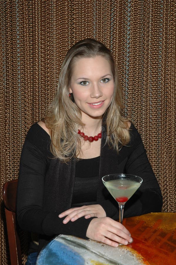 Beautiful woman sitting at a table drinking her martini. Woman looking right at the camera as if someone is approaching her at the table. Beautiful woman sitting at a table drinking her martini. Woman looking right at the camera as if someone is approaching her at the table.