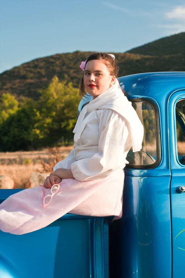 A multicultural 15 year old teen dressed in a pink poodle skirt, white blouse, sweater tied around neck, pink pearls, and pink hair ribbon is sitting in the back of a 1953 classic Chevy truck with a bright blue paint job. She is smiling. She is holding 1950s style pink eyeglasses. Her bare feet rest on the wood in the back of the truck. A multicultural 15 year old teen dressed in a pink poodle skirt, white blouse, sweater tied around neck, pink pearls, and pink hair ribbon is sitting in the back of a 1953 classic Chevy truck with a bright blue paint job. She is smiling. She is holding 1950s style pink eyeglasses. Her bare feet rest on the wood in the back of the truck.