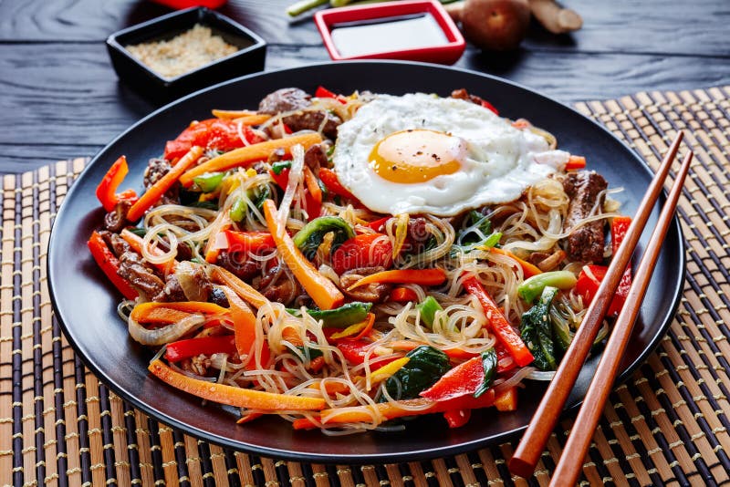 Korean japchae or chapchae stir fried noodles with vegetables: red bell pepper, carrot, shiitake mushrooms, green onion, garlic, beef, egg with dressing of sesamie seeds, oil and soy sauce, close up. Korean japchae or chapchae stir fried noodles with vegetables: red bell pepper, carrot, shiitake mushrooms, green onion, garlic, beef, egg with dressing of sesamie seeds, oil and soy sauce, close up