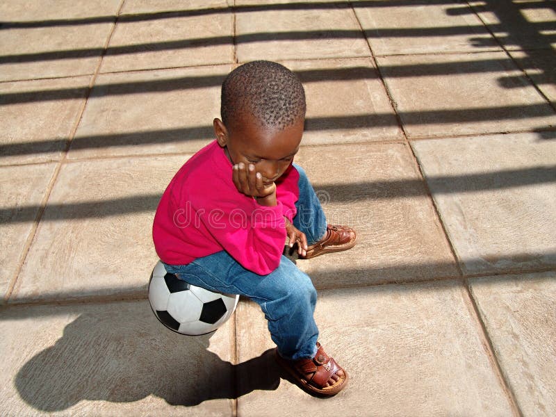 African American child sitting on the ball waiting for his friends at school for a soccer game. African American child sitting on the ball waiting for his friends at school for a soccer game.