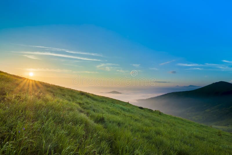 Ανατολή πέρα από τους λόφους στα βουνά με τα πράσινα WI χλόης και μπλε ουρανού