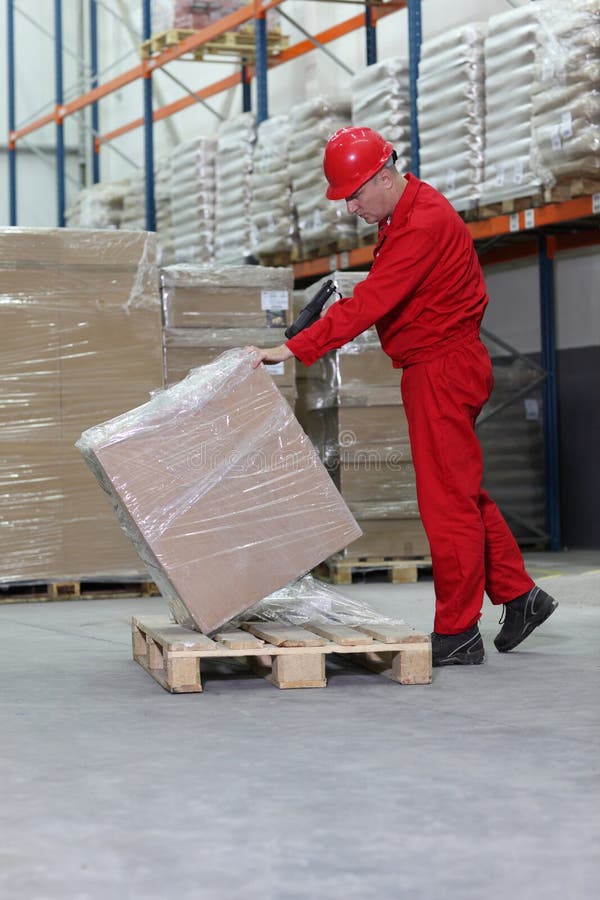 Middle aged workman in hard hat lifting cellophane wrapped box on wooden pallet in warehouse, stacked goods in background. Middle aged workman in hard hat lifting cellophane wrapped box on wooden pallet in warehouse, stacked goods in background.