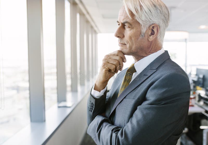 Side view shot of mature businessman standing beside office window looking outside and thinking with hand on chin. Senior professional thinking for solutions. Side view shot of mature businessman standing beside office window looking outside and thinking with hand on chin. Senior professional thinking for solutions.