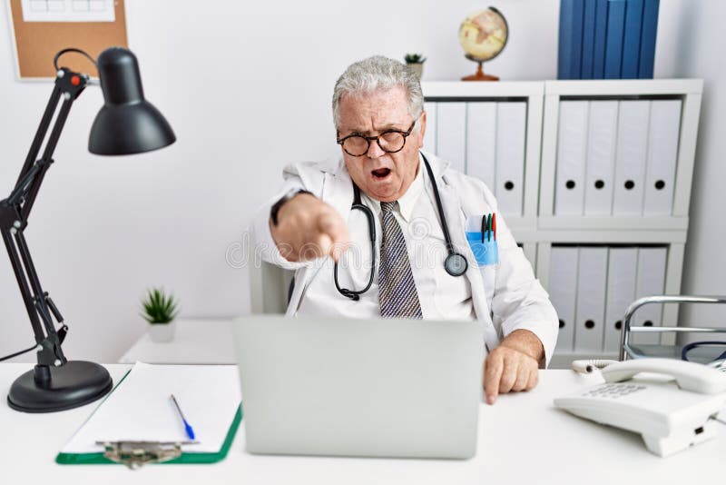 Senior caucasian man wearing doctor uniform and stethoscope at the clinic pointing displeased and frustrated to the camera, angry and furious with you. Senior caucasian man wearing doctor uniform and stethoscope at the clinic pointing displeased and frustrated to the camera, angry and furious with you