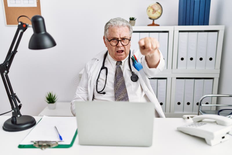 Senior caucasian man wearing doctor uniform and stethoscope at the clinic pointing displeased and frustrated to the camera, angry and furious with you. Senior caucasian man wearing doctor uniform and stethoscope at the clinic pointing displeased and frustrated to the camera, angry and furious with you