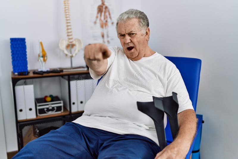 Senior caucasian man at physiotherapy clinic holding crutches pointing displeased and frustrated to the camera, angry and furious with you. Senior caucasian man at physiotherapy clinic holding crutches pointing displeased and frustrated to the camera, angry and furious with you