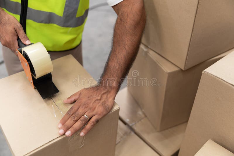 Αρσενικό κουτί από χαρτόνι συσκευασίας εργαζομένων με το διανομέα πυροβόλων όπλων ταινιών στην αποθήκη εμπορευμάτων στοκ εικόνες