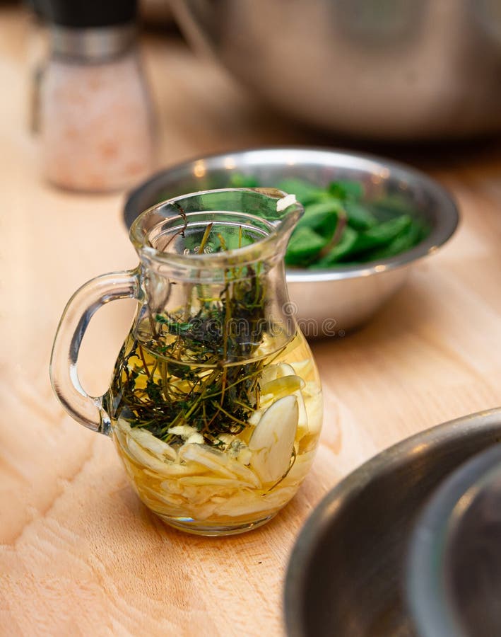 A glass jug filled with olive oil infused with fresh herbs, garlic, and lemon slices, set on a wooden kitchen countertop. A glass jug filled with olive oil infused with fresh herbs, garlic, and lemon slices, set on a wooden kitchen countertop