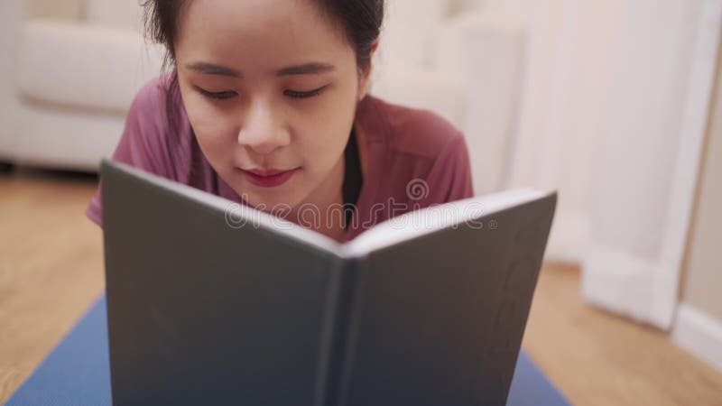 ασιάτισσα νεαρή γυναίκα ξαπλωμένη κάτω στο στρώμα γιόγκα διαβάζει βιβλίο στο σαλόνι καραντίνα covid19 μετά το σπίτι γυμναστική χαλ