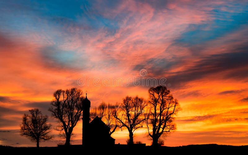 Early morning sunrise dawn with black silhouette of small church or chapel and bare trees on colorful cloudy flaming sky background. Early morning sunrise dawn with black silhouette of small church or chapel and bare trees on colorful cloudy flaming sky background