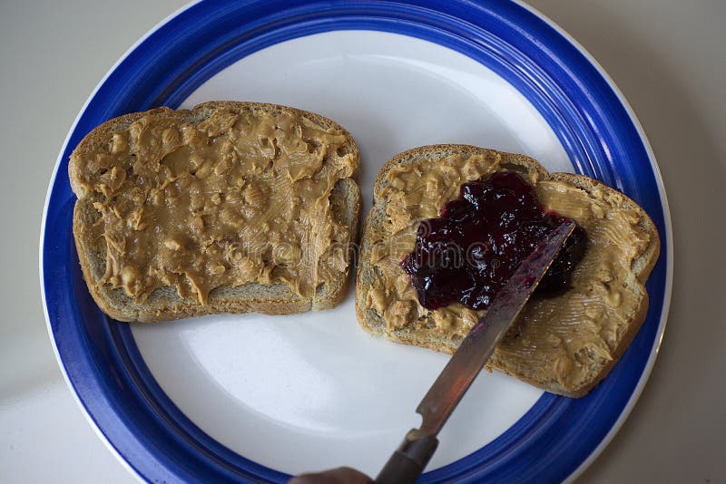 Peanut butter and jelly sandwich with 2 layers of chunky peanut butter and 1 layer of grape jelly being prepared by spreading ingredients with a butter knife. Peanut butter and jelly sandwich with 2 layers of chunky peanut butter and 1 layer of grape jelly being prepared by spreading ingredients with a butter knife.