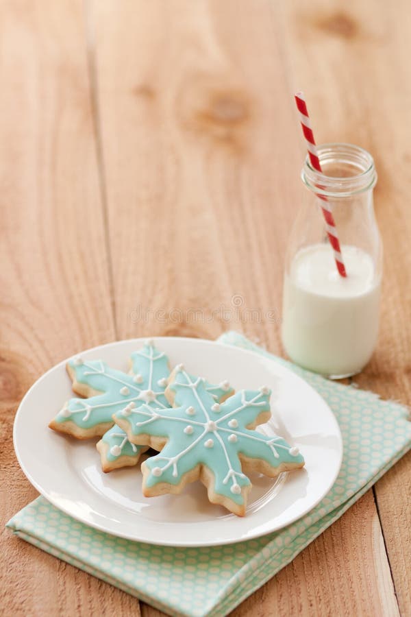 Two blue snowflake sugar cookies rest on white plate next to a small farm bottle of milk with a red and white striped straw. Two blue snowflake sugar cookies rest on white plate next to a small farm bottle of milk with a red and white striped straw.