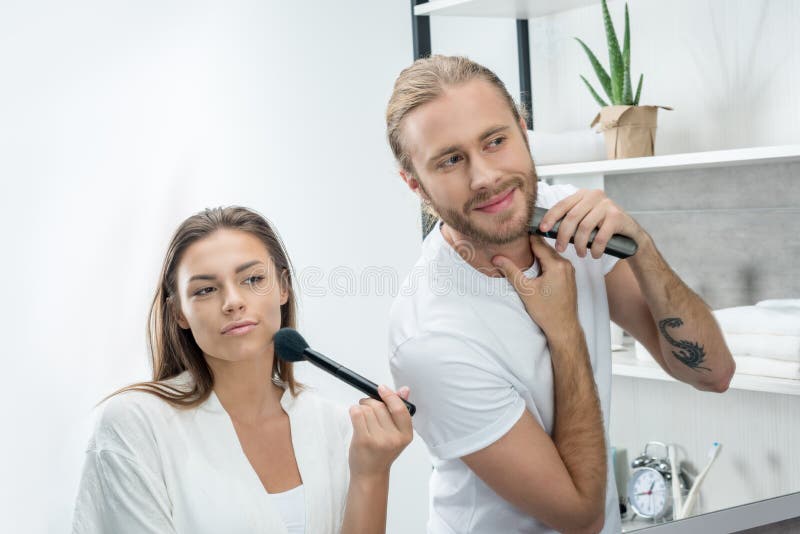 Γενειάδα ξυρίσματος ατόμων με trimmer ενώ σύζυγος που εφαρμόζει τη σκόνη προσώπου στοκ φωτογραφία Γενειάδα ξυρίσματος ατόμων με trimmer ενώ σύζυγος που εφαρμόζει τη σκόνη προσώπου στοκ φωτογραφία