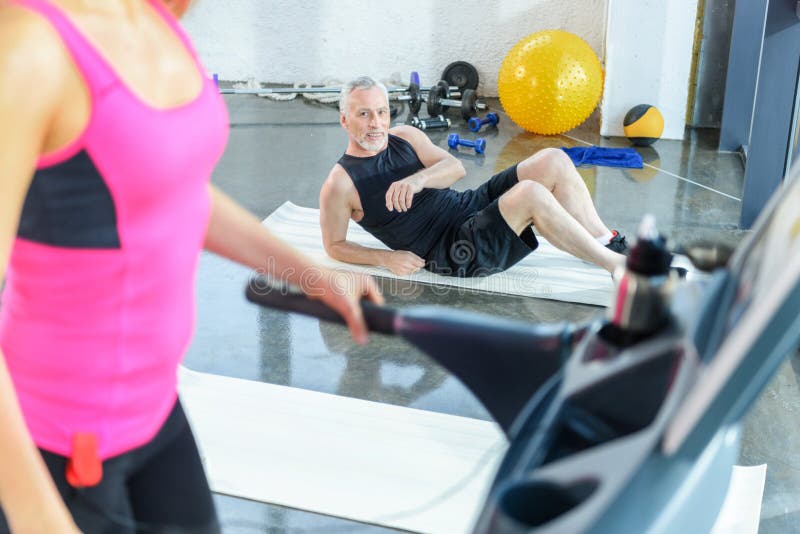 Γενειοφόρος αθλητικός τύπος που βρίσκεται στο χαλί και που εξετάζει την άσκηση γυναικών treadmill στοκ φωτογραφία Γενειοφόρος αθλητικός τύπος που βρίσκεται στο χαλί και που εξετάζει την άσκηση γυναικών treadmill στοκ φωτογραφία