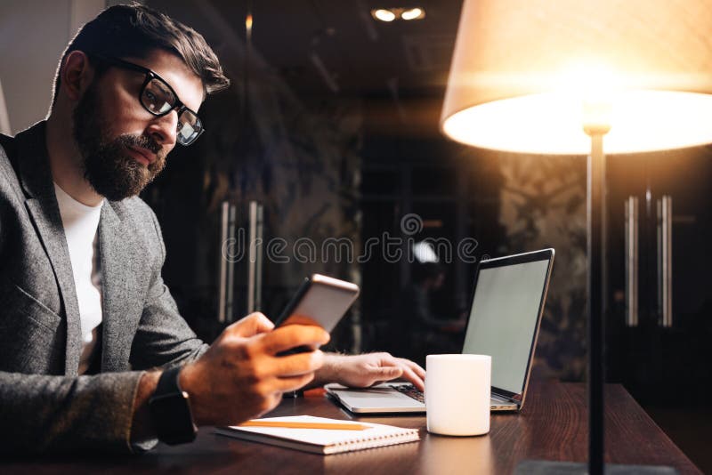 Γενειοφόρος επιχειρηματίας με το lap-top που χρησιμοποιεί το τηλέφωνο κυττάρων στο γραφείο σοφιτών νύχτας Κείμενο δακτυλογράφησης