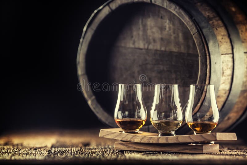 Glencairn whiskey tasting cups on a wooden serving, with a whisky barrel in the dark background. Glencairn whiskey tasting cups on a wooden serving, with a whisky barrel in the dark background.