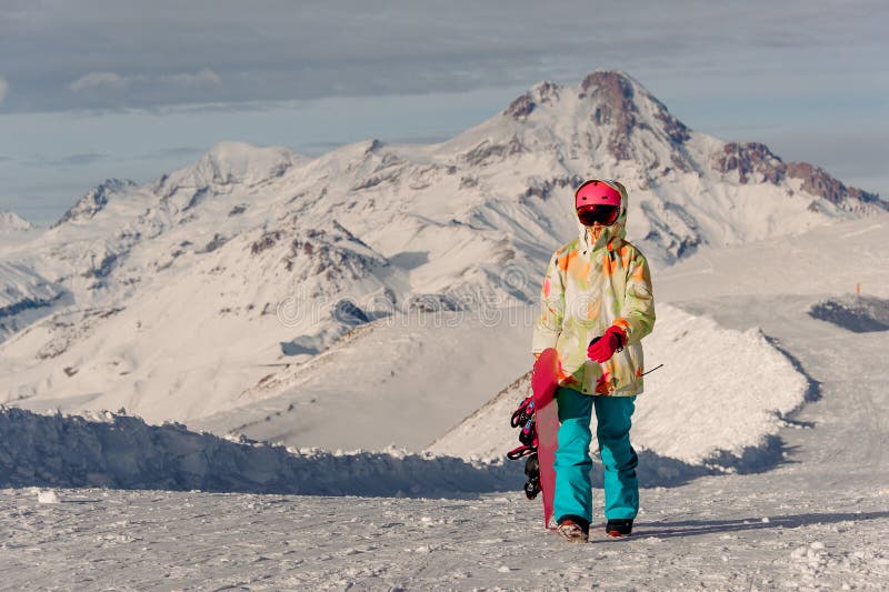 Γυναίκα snowboarder στο ρόδινο αθλητικό κράνος και φωτεινό ζωηρόχρωμο sportswear ενάντια στις αιχμές βουνών στοκ εικόνα Γυναίκα snowboarder στο ρόδινο αθλητικό κράνος και φωτεινό ζωηρόχρωμο sportswear ενάντια στις αιχμές βουνών στοκ εικόνα