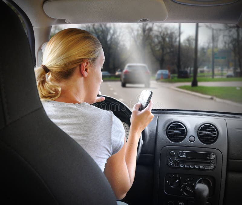 A young woman is on the cell phone texting and driving with a road in the windshield for an danger or distracted driving concept. A young woman is on the cell phone texting and driving with a road in the windshield for an danger or distracted driving concept.