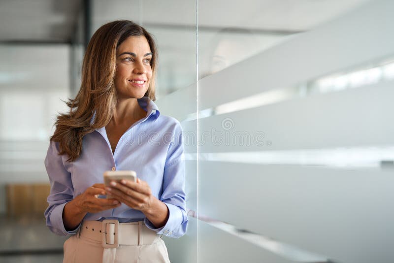 Smiling middle aged woman business investor using mobile banking apps on cellular phone standing in office. Mature businesswoman executive holding cellphone working on smartphone looking away. Smiling middle aged woman business investor using mobile banking apps on cellular phone standing in office. Mature businesswoman executive holding cellphone working on smartphone looking away.