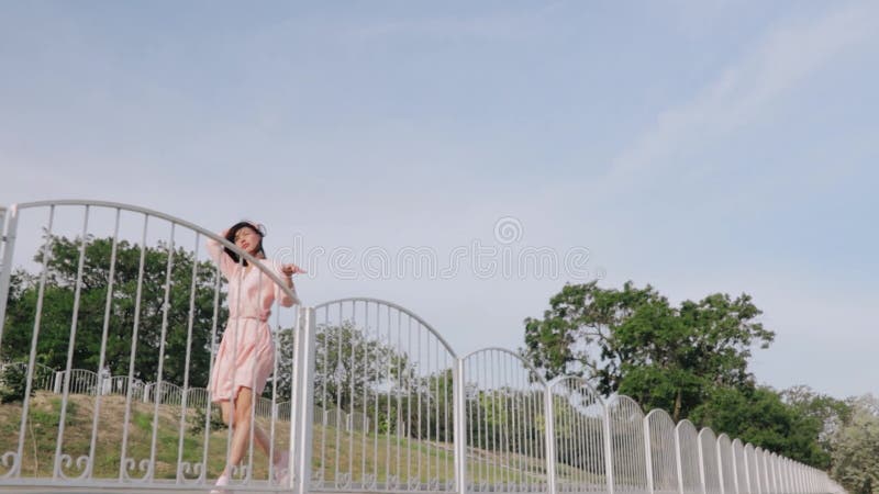 γυναίκα με ροζ φόρεμα χαμογελώντας περπατώντας στην όχθη του ποταμού με μαλλιά να πετούν στο μπλε φόντο του ανέμου