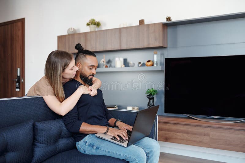 Pretty young women hugging her boyfriend from behind when he is working on laptop when sitting on sofa at home. Pretty young women hugging her boyfriend from behind when he is working on laptop when sitting on sofa at home