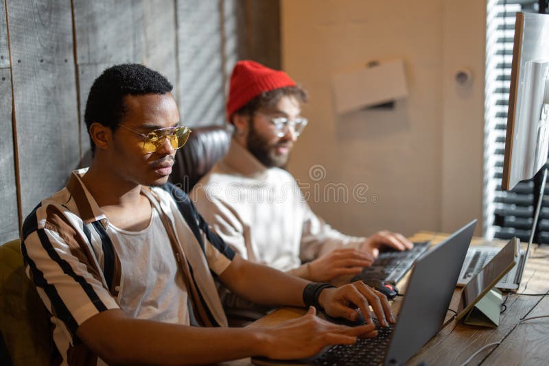 Two men with different nationality working on computers, sitting together at cozy home office. Concept of freelance and remote work. Stylish male hipsters programming together. Two men with different nationality working on computers, sitting together at cozy home office. Concept of freelance and remote work. Stylish male hipsters programming together