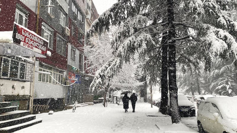 21022025 Μπατούμι Γεωργία : Δύο άνθρωποι περπατάνε κάτω από μια ομπρέλα σε ένα χιονισμένο πεζοδρόμιο κατά τη διάρκεια έντονης χιονόπτωσης περικυκλωμένοι από δέντρα κτιρίων και σταθμευμένα αυτοκίνητα σε μια χειμερινή αστική σκηνή