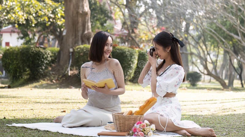 Two attractive young Asian women enjoying picnic in the park together, taking a photo from camera, reading a book. Two attractive young Asian women enjoying picnic in the park together, taking a photo from camera, reading a book.