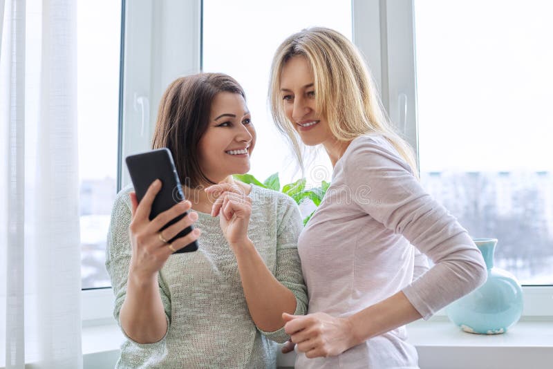 Two middle-aged women friends looking together at smartphone screen. Females at home near window in winter season. Relationship, lifestyle, leisure, friendship, technology, mature people concept. Two middle-aged women friends looking together at smartphone screen. Females at home near window in winter season. Relationship, lifestyle, leisure, friendship, technology, mature people concept