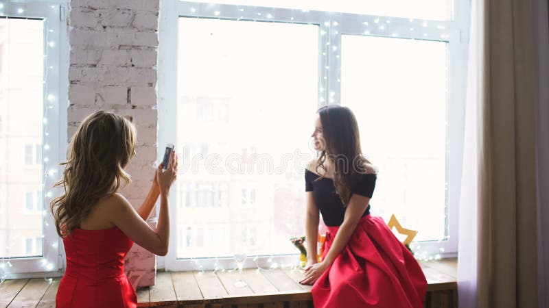 Δύο νέες γυναίκες στο κόκκινο κοκτέιλ ντύνουν την τοποθέτηση και photograps στο smartphone στο παράθυρο