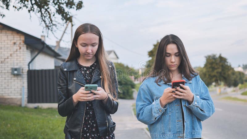 Two girlfriends watch a smartphone and walk through the village. Two girlfriends watch a smartphone and walk through the village