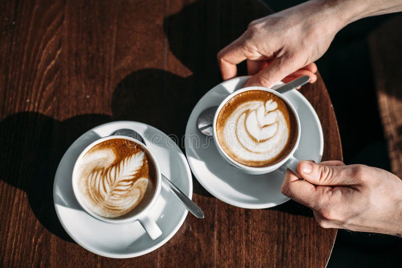 Δύο φλυτζάνια του cappuccino με την τέχνη latte στον ξύλινο πίνακα στα χέρια του ατόμου στοκ εικόνες με δικαίωμα ελεύθερης χρήσης Δύο φλυτζάνια του cappuccino με την τέχνη latte στον ξύλινο πίνακα στα χέρια του ατόμου στοκ εικόνες με δικαίωμα ελεύθερης χρήσης