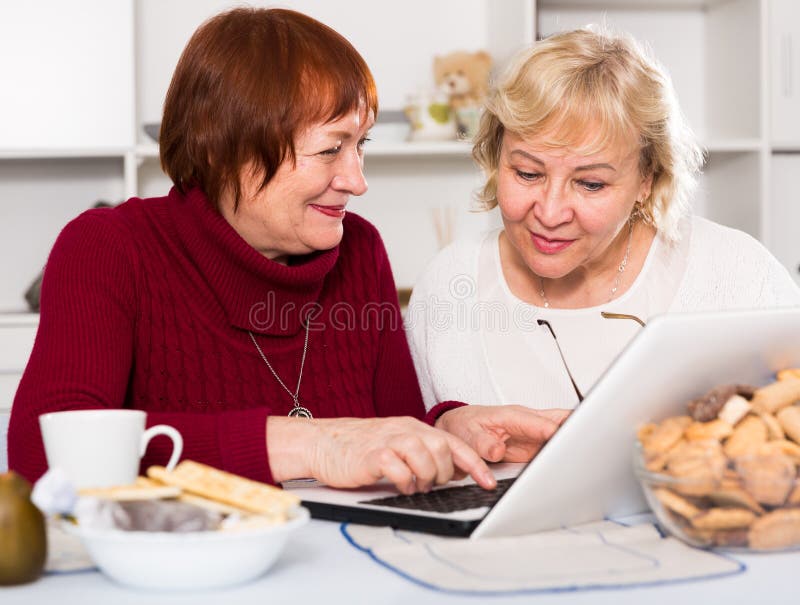 Two smiling elderly women enjoying time at home, drinking tea and using laptop. Two smiling elderly women enjoying time at home, drinking tea and using laptop