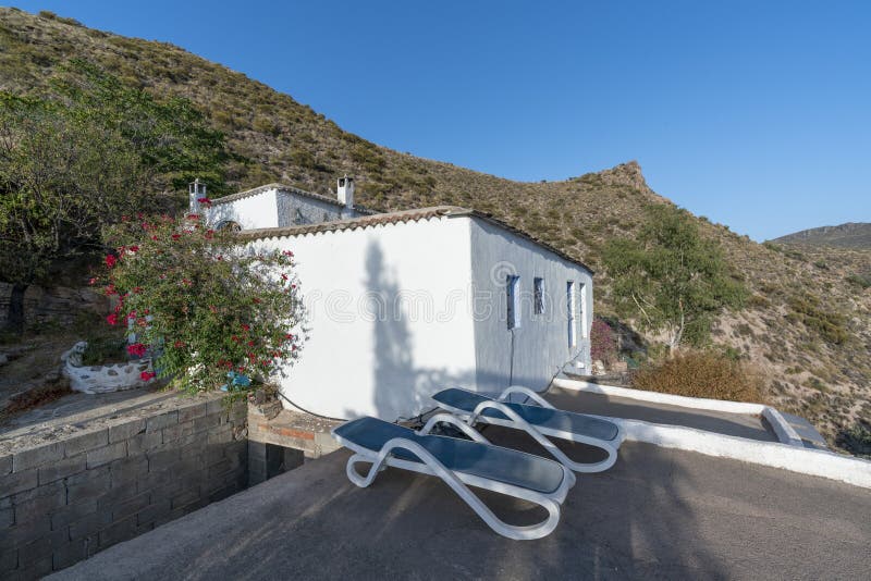 Abandoned farmhouse, the facade is white and has windows and doors, on the terrace there are 2 deck chairs, behind there are trees and mountains, and the sky is clear. Abandoned farmhouse, the facade is white and has windows and doors, on the terrace there are 2 deck chairs, behind there are trees and mountains, and the sky is clear