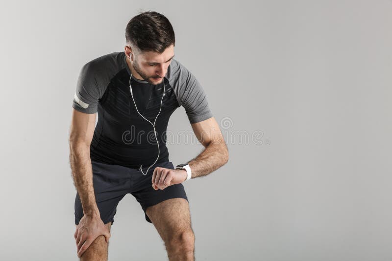 Image of young tired man in sportswear using smartwatch and earphones while working out isolated over gray background. Image of young tired man in sportswear using smartwatch and earphones while working out isolated over gray background
