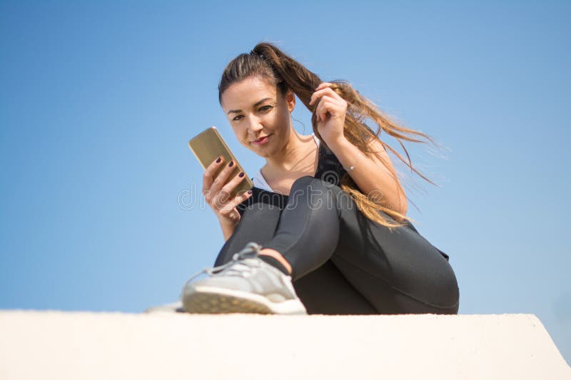 Low angle view of beautiful young fit woman in sportswear using mobile phone against blue sky. Low angle view of beautiful young fit woman in sportswear using mobile phone against blue sky.