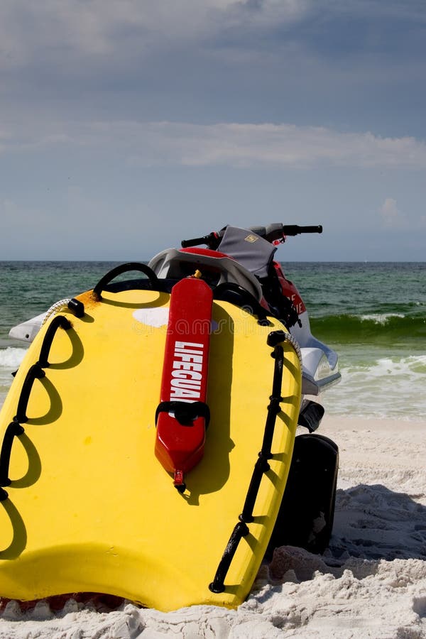 Bright yellow lifesaving equipment attached to the back of a water craft and a red flotation device with the word lifeguard in white and the beach in the background. Bright yellow lifesaving equipment attached to the back of a water craft and a red flotation device with the word lifeguard in white and the beach in the background.