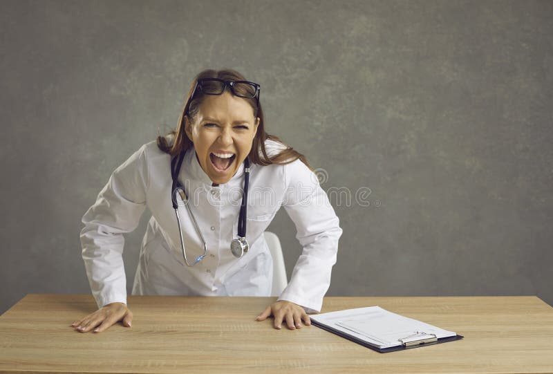 Angry furious female doctor shouting at camera standing behind desk studio shot. Frustrated woman medical worker in white coat and stethoscope showing emotional rage and aggressive expression. Angry furious female doctor shouting at camera standing behind desk studio shot. Frustrated woman medical worker in white coat and stethoscope showing emotional rage and aggressive expression